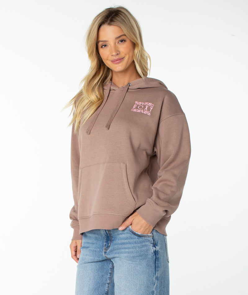 A woman with long blonde hair wears a light brown Roxy Wailea Hoodie featuring a pink design on the chest and blue jeans, standing against a plain white background.