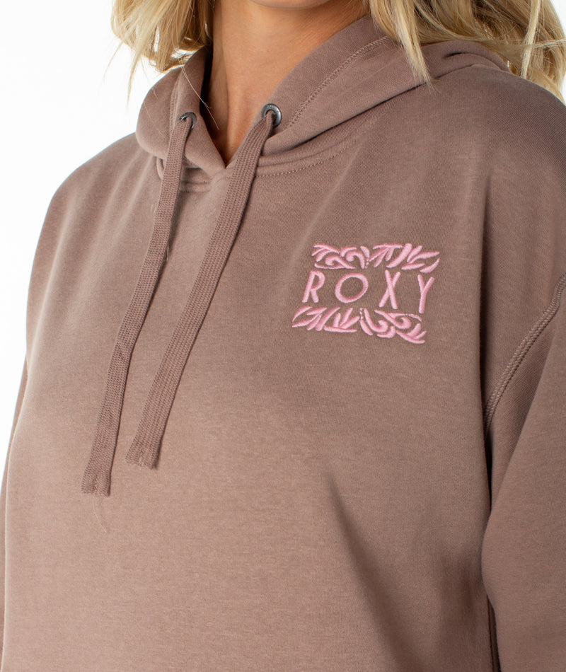 A person is shown wearing the Roxy Wailea Hoodie—a brown, ultra-soft hoodie featuring a pink ROXY logo and decorative border on the left chest. The image focuses on the lower face, neck, and upper torso, highlighting the effortless comfort of this signature piece.