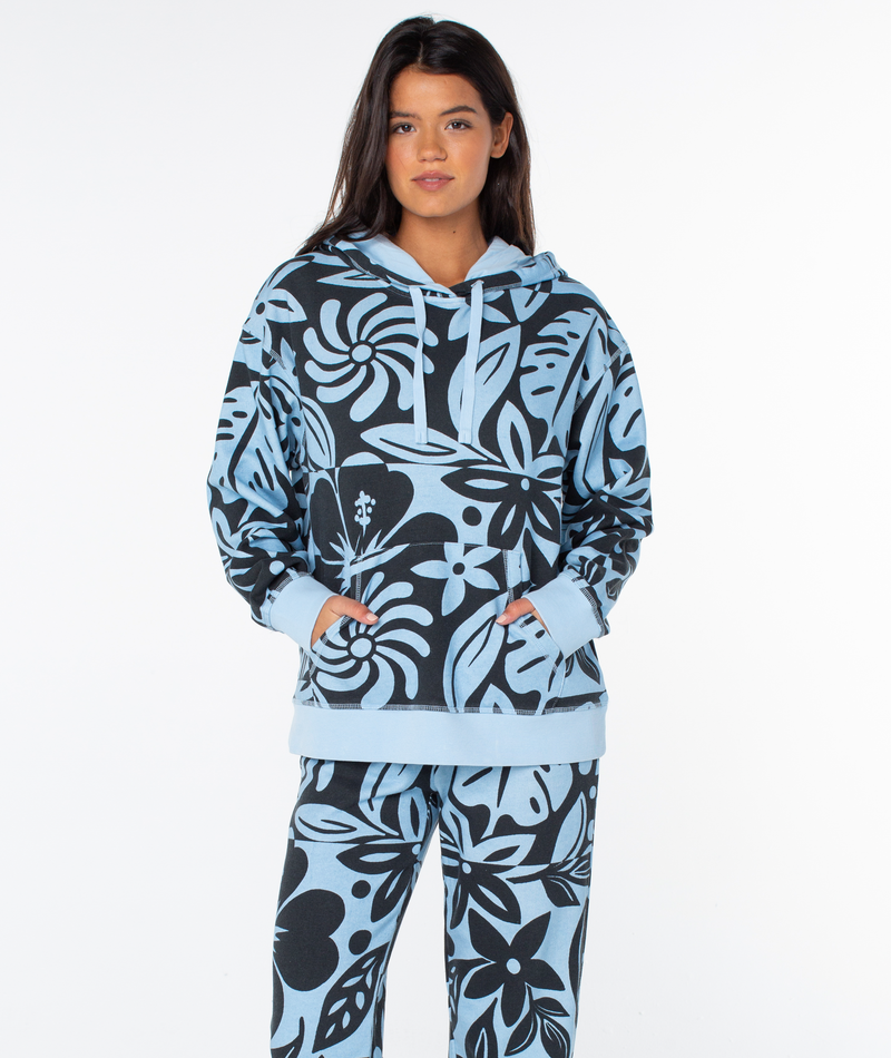 A person wears a light blue Roxy Maile Printed Hoodie and matching pants featuring a dark blue tropical floral print and a front kangaroo pocket.