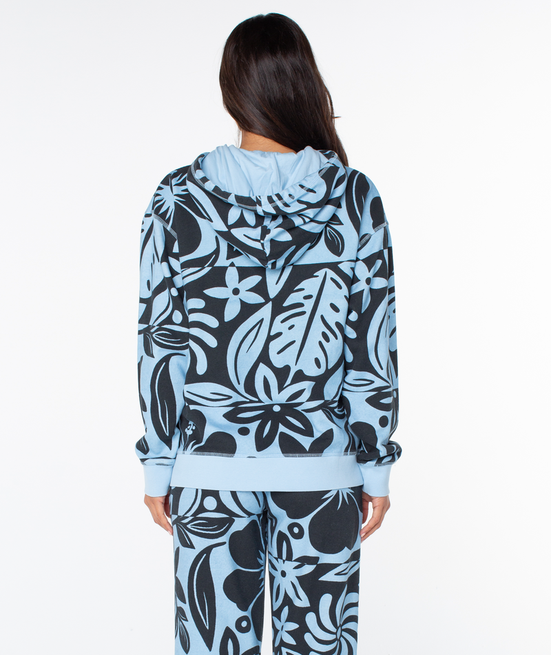 Back view of a person wearing a Roxy Maile Printed Hoodie and matching pants, both featuring an oversized light blue fit with a bold black floral and leaf print with beachy graphics.