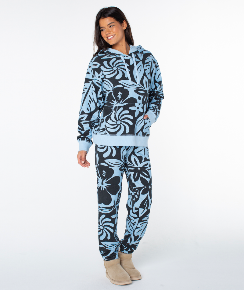 A smiling woman wearing a Cerulean Cool Surf Roxy Maile Printed Hoodie and matching sweatpants with a dark blue floral and tropical leaf print.