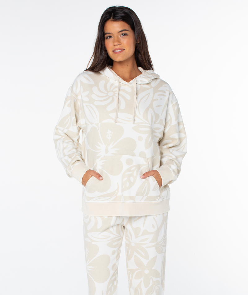 A woman wearing a Roxy Maile Printed Hoodie in an oversized, beige and cream hibiscus print with a front kangaroo pocket and matching lounge pants.