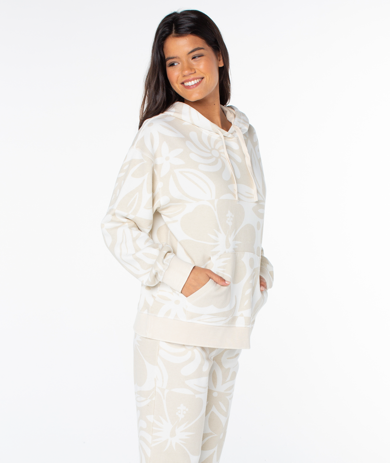 A smiling woman wears a cream-colored Roxy Maile Printed Hoodie, featuring an oversized fit, a tonal hibiscus flower print, and a front kangaroo pocket.