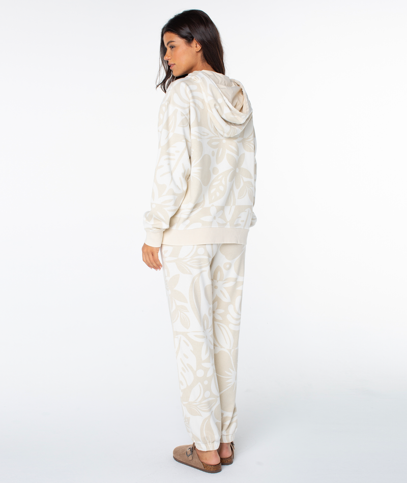 A woman wearing a Roxy Maile Printed Hoodie and matching sweatpants in cream, both featuring a large tonal leaf print with beachy graphics.
