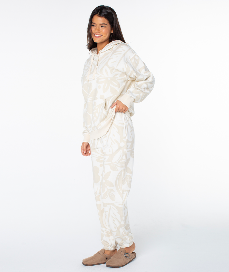 A smiling woman wears an oversized Maile Printed Hoodie and matching joggers in Parchment Cool Surf with a light-colored tropical leaf print, paired with tan slip-on shoes.