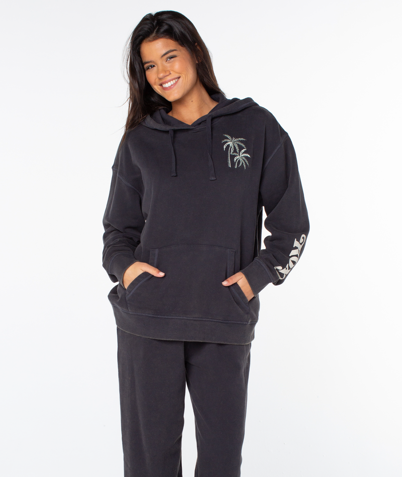 A woman smiles in the Roxy Maile Hoodie, a relaxed fit with palm tree embroidery and front pocket, paired with matching black pants against a plain white background.