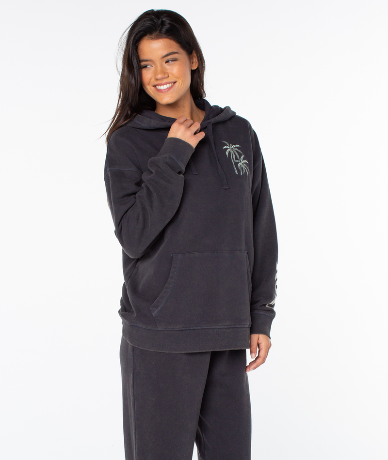 A woman with long, dark hair smiles in a relaxed fit Maile Hoodie by Roxy, featuring a small palm tree graphic, paired with matching sweatpants against a plain white background.
