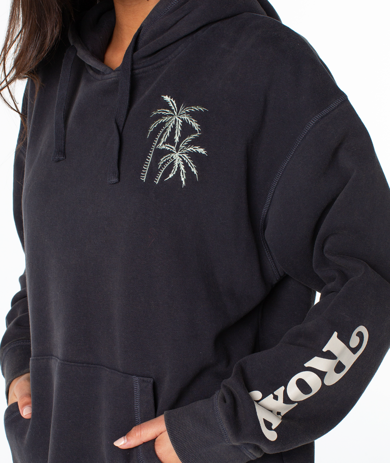 Person wearing the Maile Hoodie by Roxy—a black cotton blend hoodie with two embroidered palm trees on the chest, ROXY printed in white on the sleeve, relaxed fit, and hands in the front pocket.