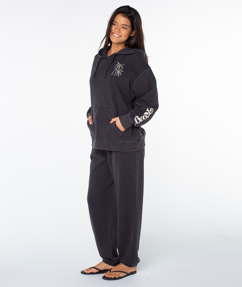 A woman wears the Roxy Maile Hoodie in Phantom Palm Tree Resort, paired with black pants and flip-flops, smiling with her hands in her hoodie pocket against a plain white background.