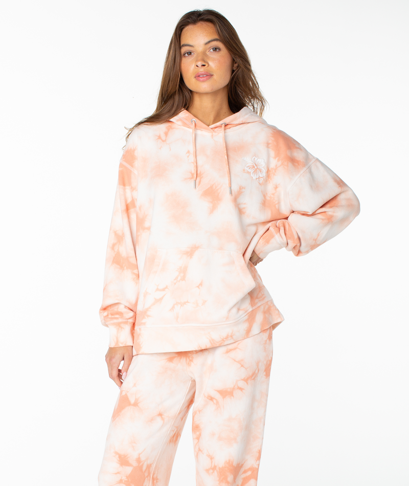 A woman wears the Roxy Maile Tie Dye Hoodie in Coral Pink Surf Day Tie Dye Wash with matching sweatpants, standing confidently with one hand on her hip against a plain white background—perfect loungewear for relaxing in style.