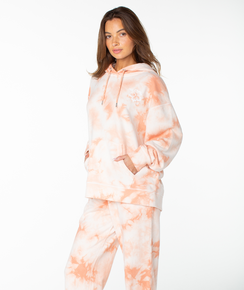 A woman stands with her hands in her pockets, wearing the Roxy Maile Tie Dye Hoodie—a relaxed, peach and white cotton sweatshirt—paired with matching sweatpants against a plain white background, embodying effortless loungewear style.