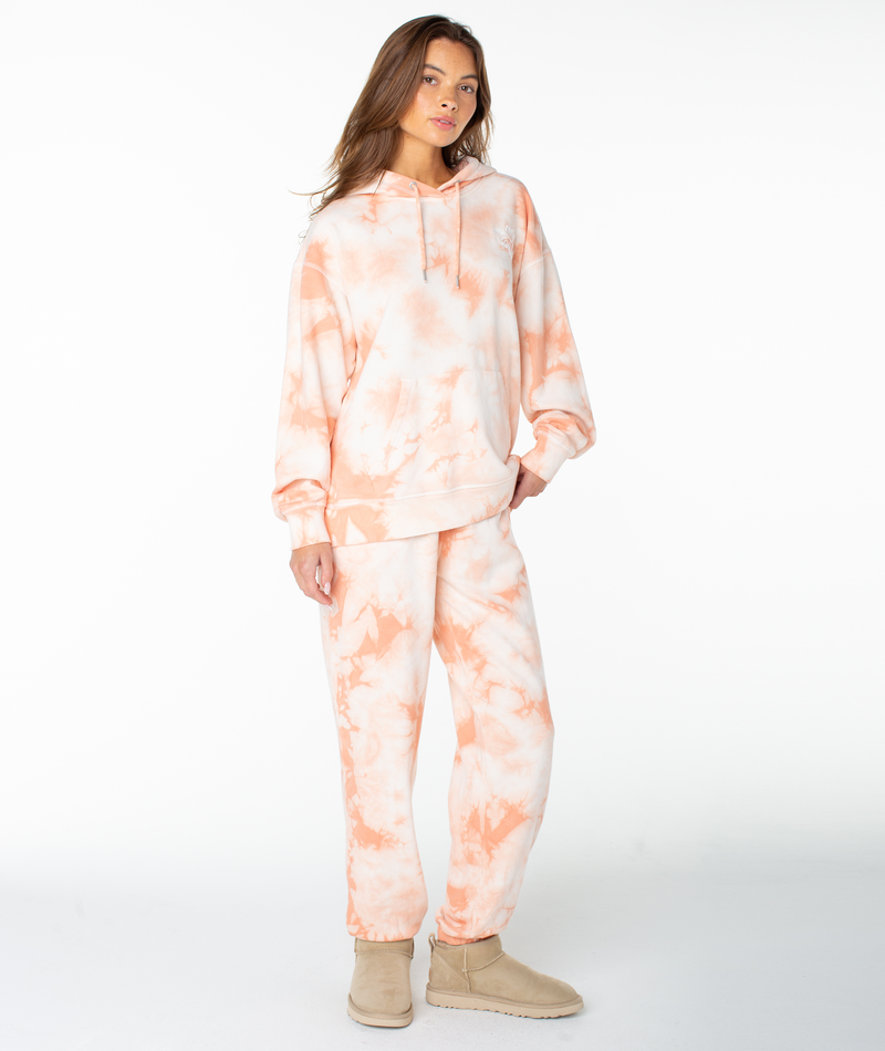 A woman wearing the Roxy Maile Tie Dye Hoodie and matching sweatpants, styled as cozy loungewear with beige boots, stands against a plain white background.