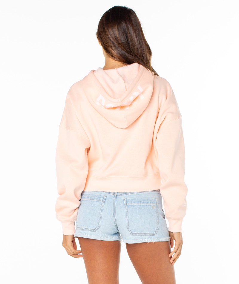 A woman stands facing away from the camera against a white background, wearing a light pink Roxy Breakwater Hoodie in a cropped fit and light blue denim shorts.