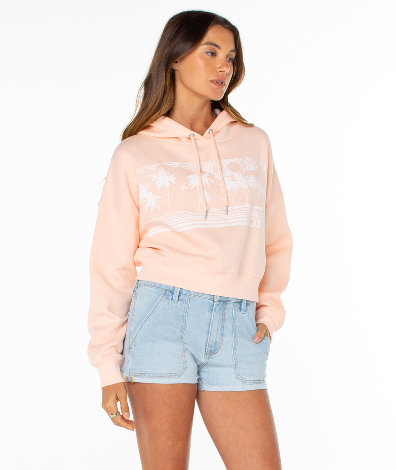 A woman models the Roxy Breakwater Hoodie in light pink, featuring a white palm tree graphic and cropped fit, paired with light blue denim shorts. She poses against a white background with one hand in her pocket, looking to the side.