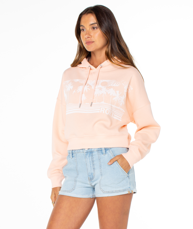 A woman wears the Roxy Breakwater Hoodie in light peach with a white palm tree graphic and cropped fit, styled with light blue denim shorts, set against a plain white background.