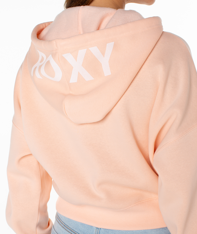 A person wears the Roxy Breakwater Hoodie—a light peach, cropped cotton blend with "ROXY" in large white letters on the hood—paired with light blue jeans.