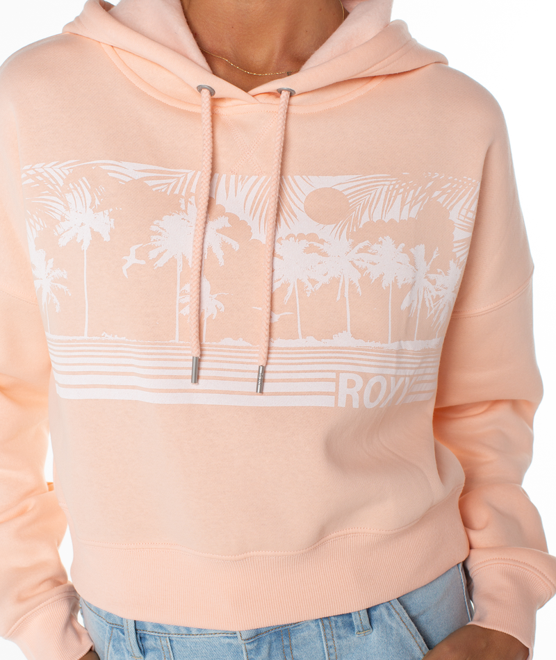 Wearing the Roxy Breakwater Hoodie in light peach with a white palm tree, sun, and stripe graphic on the chest. This relaxed cotton blend hoodie features drawstrings, a cropped fit, and pairs perfectly with light blue jeans.