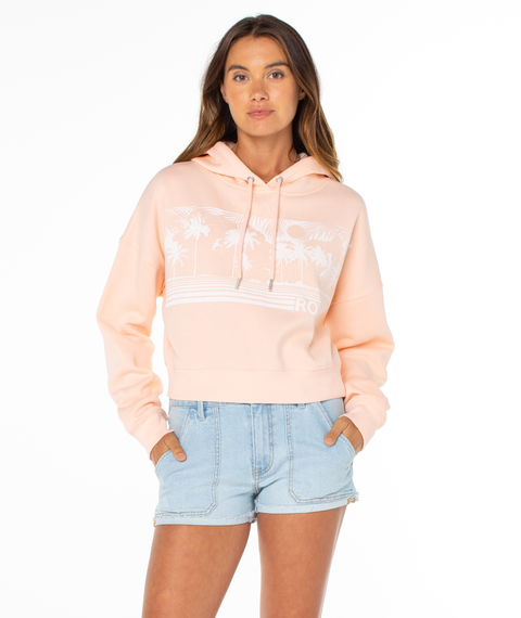 A woman stands against a white background wearing the Roxy Breakwater Hoodie, featuring a relaxed cotton blend with a white palm tree graphic, paired with light blue denim shorts. Her hands are in her pockets and she looks forward.