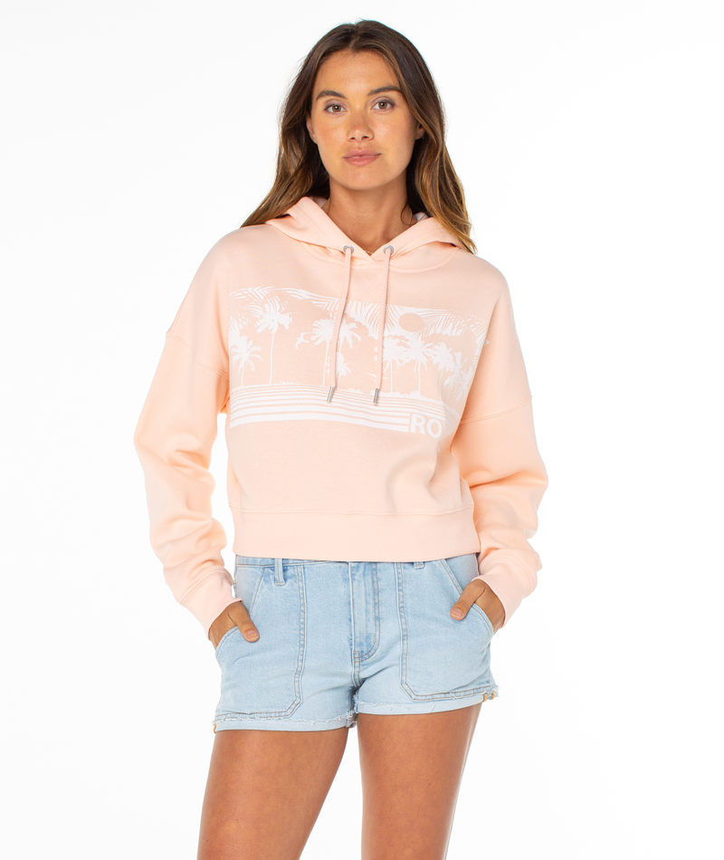 A woman stands against a white background wearing the Roxy Breakwater Hoodie, featuring a relaxed cotton blend with a white palm tree graphic, paired with light blue denim shorts. Her hands are in her pockets and she looks forward.