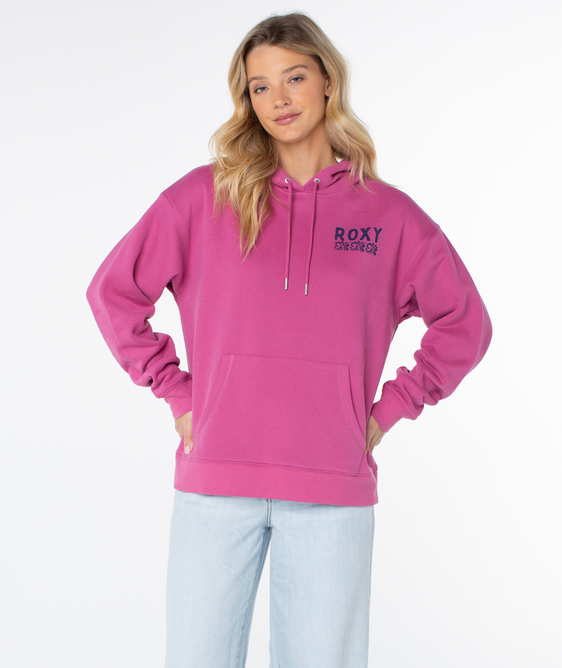 A woman wears the Raya Hoodie by Roxy in bright pink, featuring ROXY text and a chest graphic, paired with light blue jeans, standing hands on hips against a plain white background.