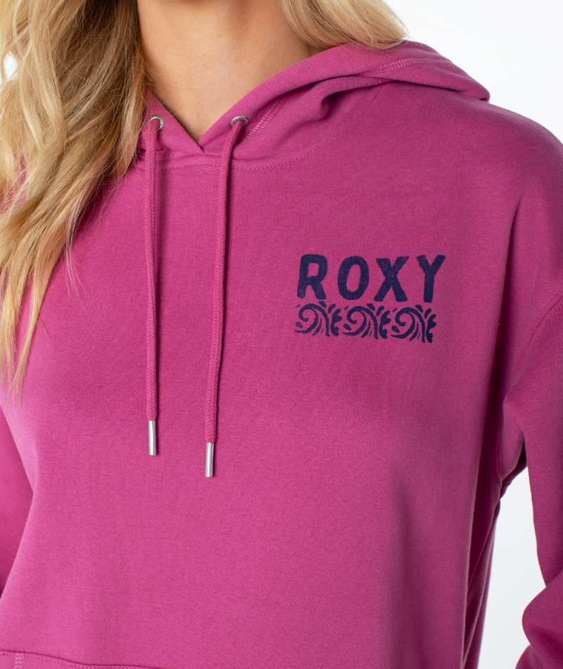 A person wears the Raya Hoodie by Roxy in magenta, featuring a black ROXY logo and decorative pattern on the left chest. Only their lower face and part of their hair show, emphasizing the graphic details.
