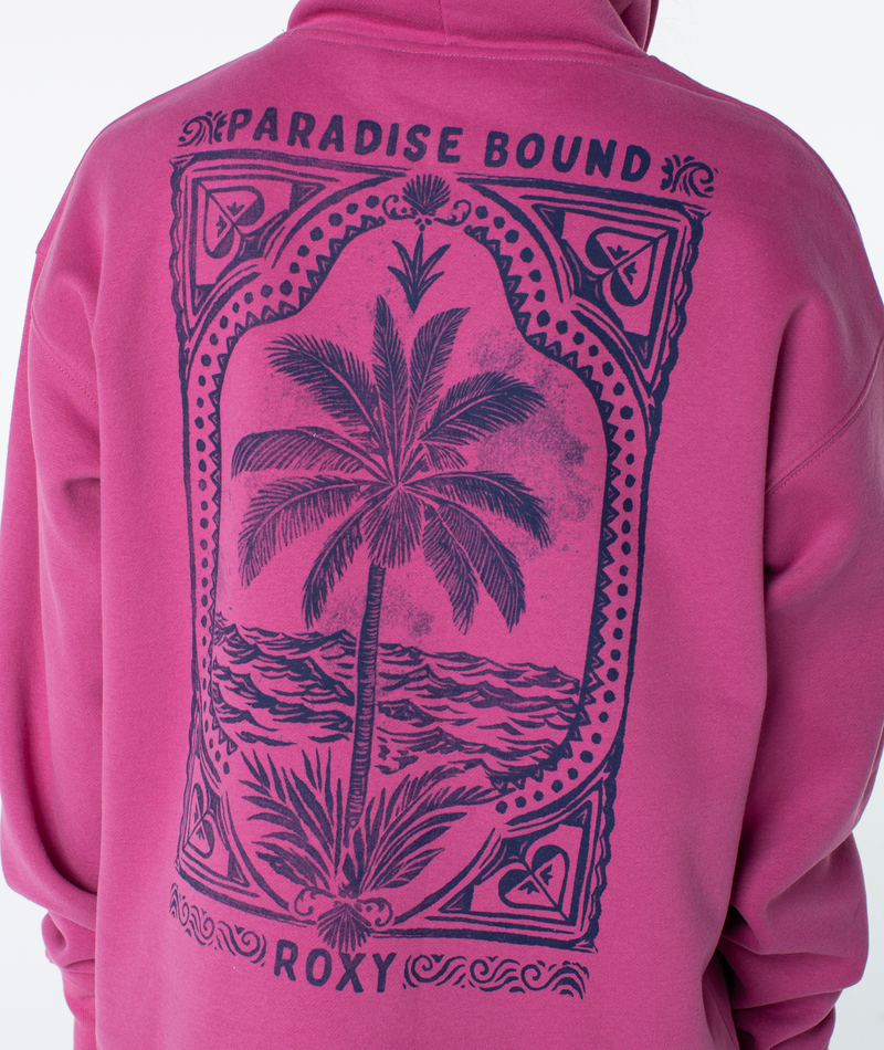 A person wearing the Roxy Raya Hoodie, an oversized sweatshirt with a large back graphic of a palm tree, ocean waves, and PARADISE BOUND ROXY text.