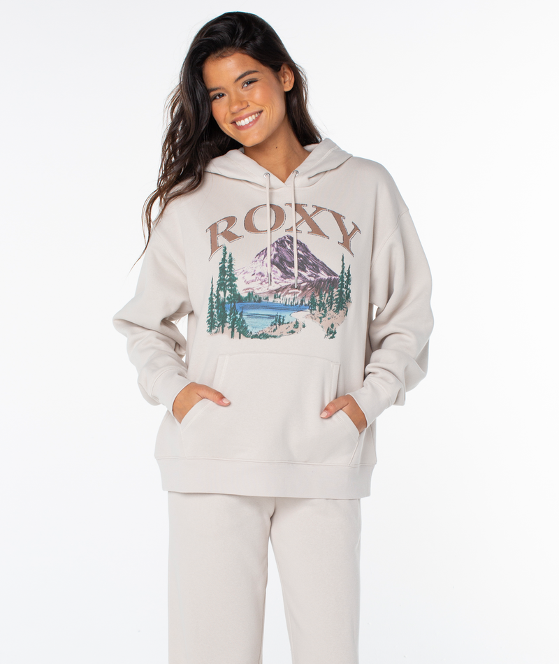 Woman smiling in an oversized fit Raya Hoodie by Roxy with a mountain and lake graphic, ROXY text, and matching pants. Hands are in hoodie pockets against a plain white background.