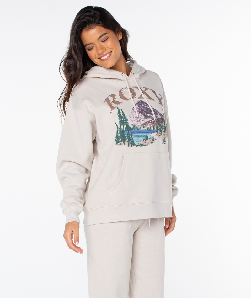 A woman in an oversized beige Raya Hoodie and sweatpants by Roxy smiles while looking down. The hoodie has a mountain, trees, lake graphic, and the word ROCKY on the front. Plain white background.