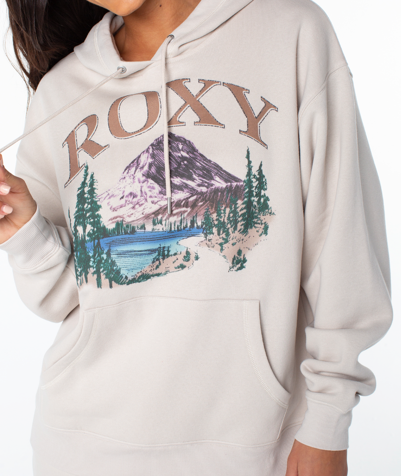 A woman wears the Roxy Raya Hoodie in beige, featuring ROXY text above a mountain, lake, and trees graphic. This oversized sweatshirt is shown with her holding a drawstring and her face partially out of frame.