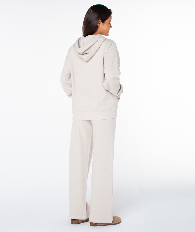 A person faces away, wearing an oversized fit Raya Hoodie by Roxy in light beige with matching wide-leg pants and tan slippers, set against a plain white background.