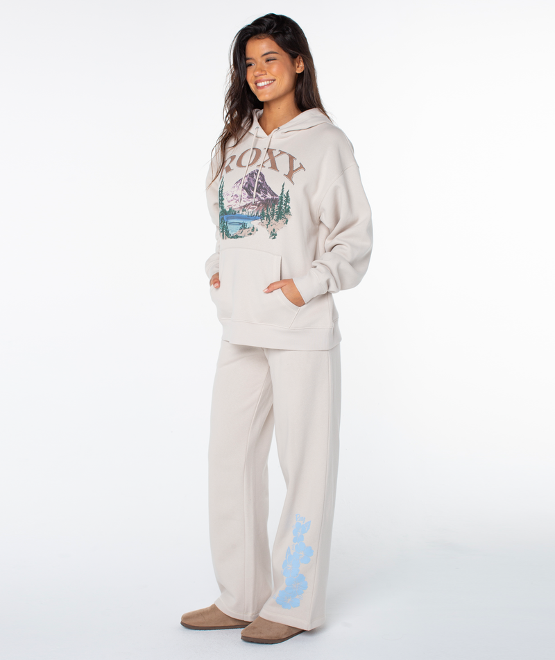 A woman smiles in the Roxy Raya Hoodie in Pumice Stone Adventures with a mountain graphic, paired with matching cream sweatpants featuring blue bears, and tan shoes against a white backdrop.