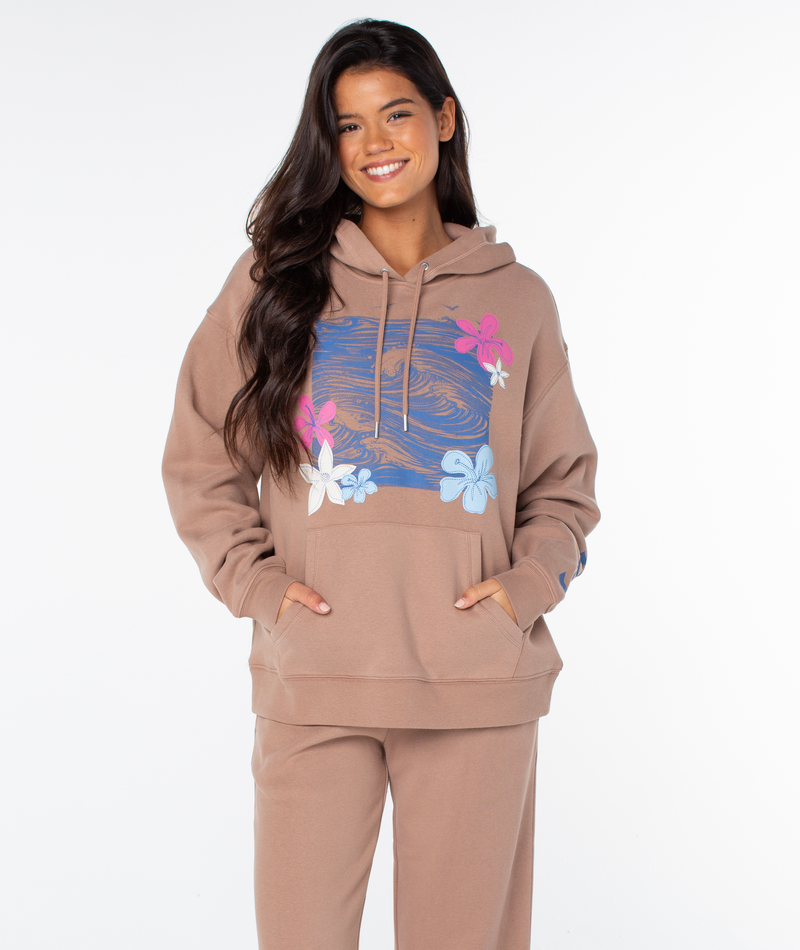 A woman wears the Roxy Raya Hoodie with matching pants, hands in pockets. The oversized hoodie features a blue rectangle with pink, white, and blue floral graphics. She smiles against a plain white background.