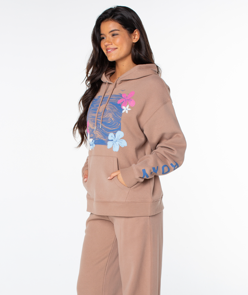 A woman with long dark hair wears the Roxy Raya Hoodie in tan with blue and pink floral graphics and sleeve lettering, smiling as she stands against a plain white background.