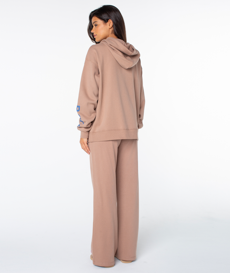 A woman stands with her back to the camera, wearing the loose-fitting Raya Hoodie by Roxy with graphic accents and matching wide-leg pants. Her hair is down, partially covering her face, against a plain white background.