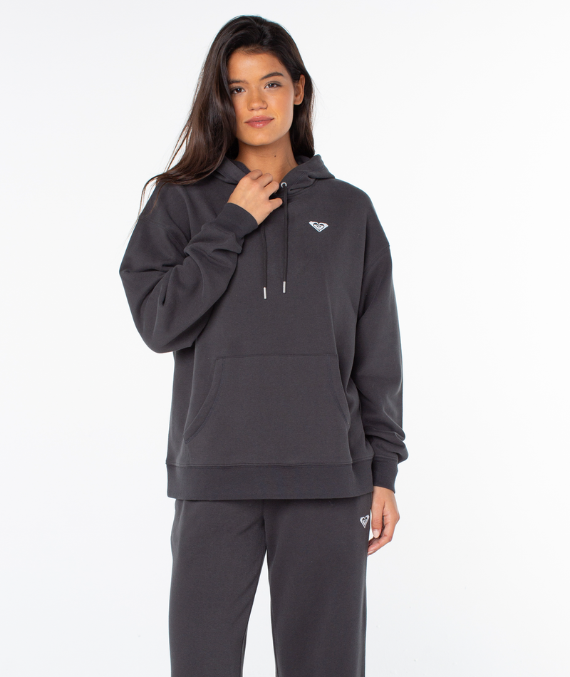 A woman wears an oversized fit dark gray Raya Hoodie by Roxy with a small white logo on the left chest, paired with matching sweatpants, standing against a white background and holding a drawstring.