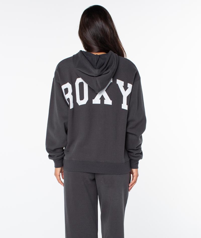 Wearing an oversized fit Raya Hoodie by Roxy, a person with long hair faces away from the camera. The graphic sweatshirt displays bold white ROXY lettering across the back.