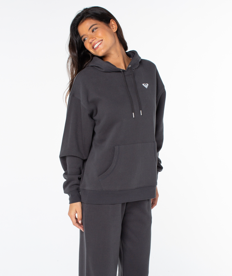 A woman smiles in an oversized fit, dark Raya Hoodie by Roxy with matching pants, standing against a plain white background, head tilted slightly and hands relaxed at her sides.
