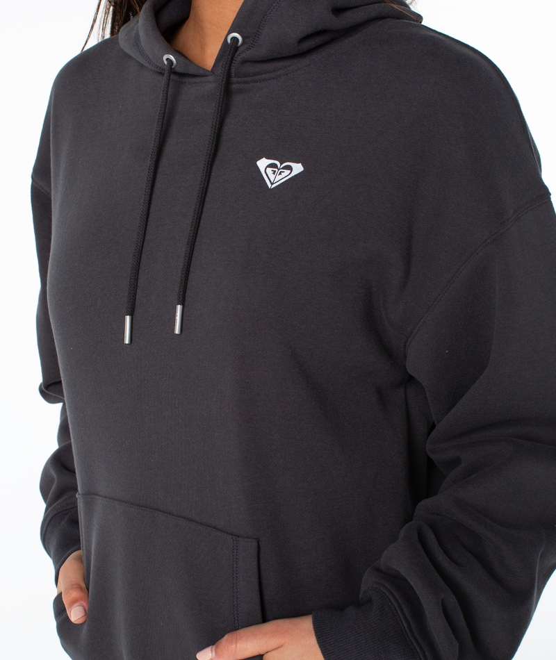 Person wears an oversized fit black Raya Hoodie by Roxy with a small white chest logo, drawstrings down, and one hand in the pocket, set against a plain white background.