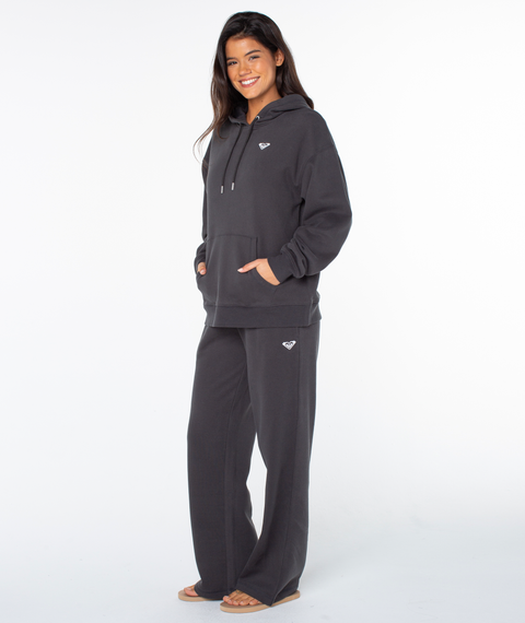 A person smiles in the Raya Hoodie by Roxy in Phantom Hey Girl, paired with loose pants and sandals, hands in pockets, showcasing an oversized fit against a plain white background.