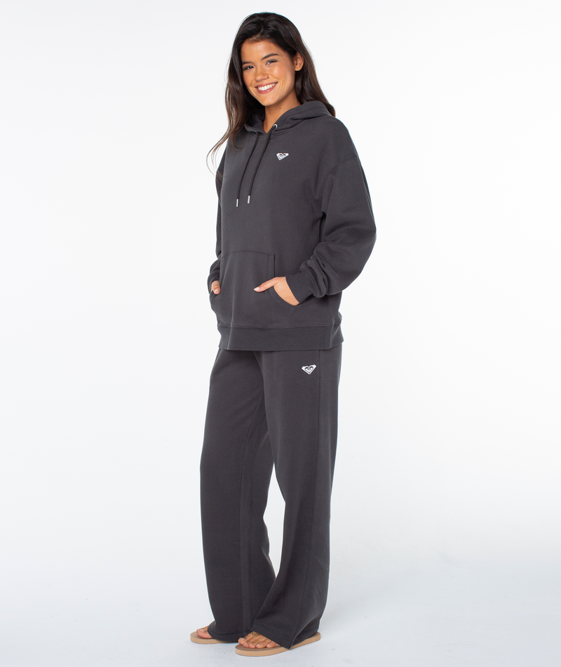 A person smiles in the Raya Hoodie by Roxy in Phantom Hey Girl, paired with loose pants and sandals, hands in pockets, showcasing an oversized fit against a plain white background.
