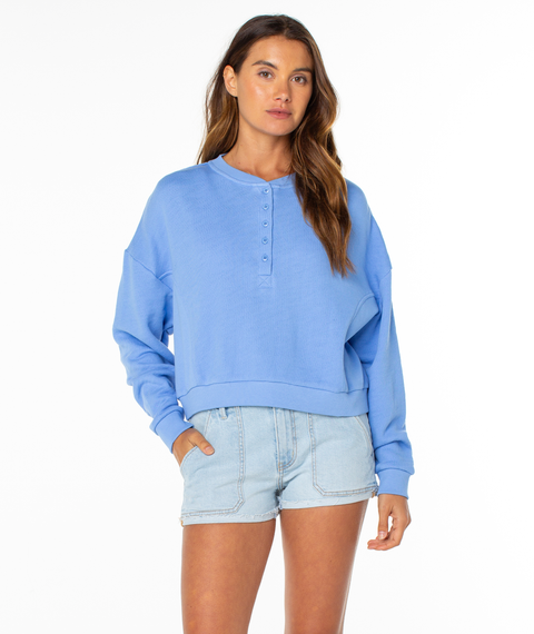A woman stands against a white background, wearing a light blue Roxy Overcast Hoodie and light denim shorts with her left hand in her pocket—a true wardrobe staple for effortless style.