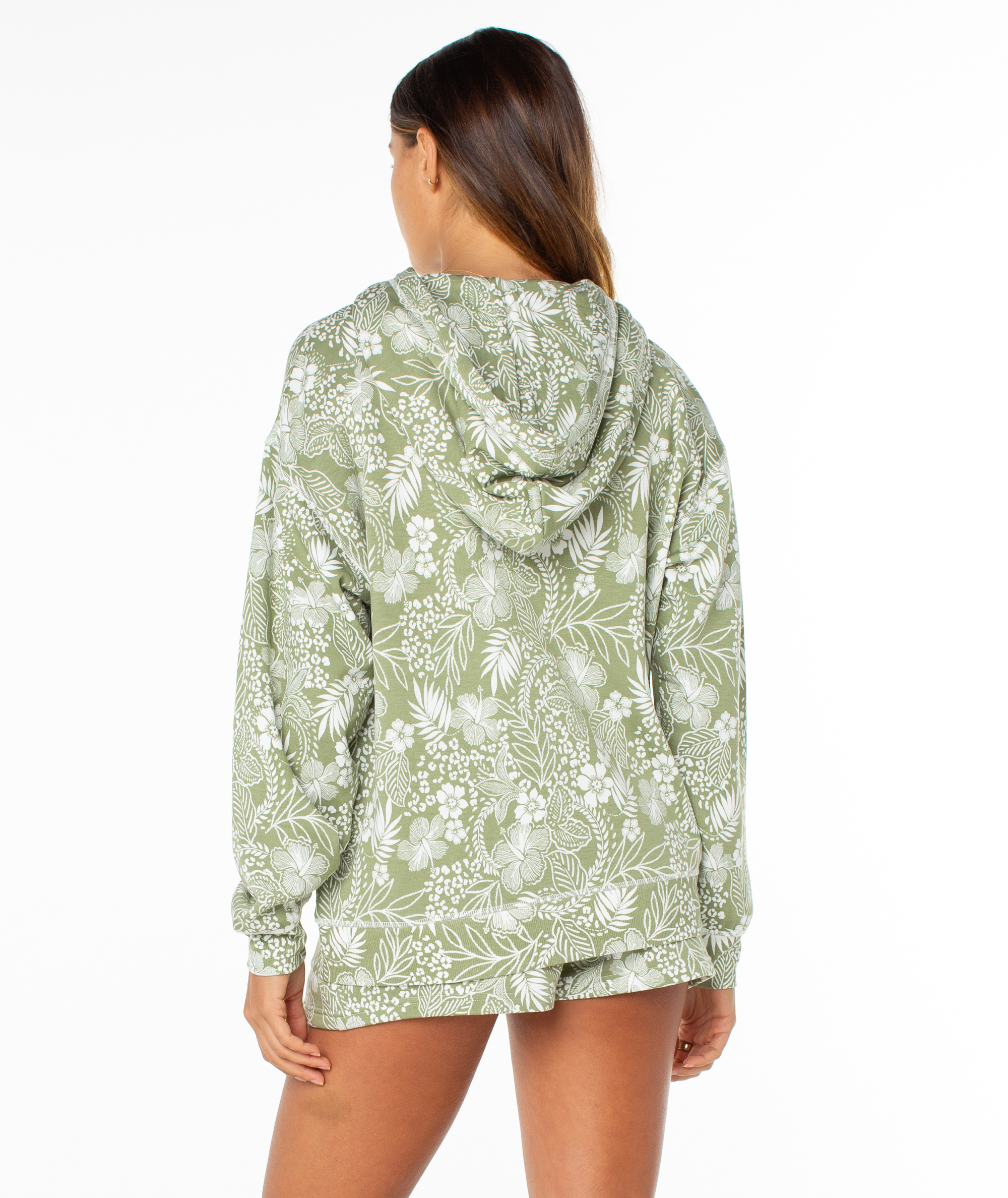 A woman with long brown hair, seen from behind, wears the Roxy Surfing By Moonlight Printed Hoodie in light green with a white tropical floral print, made from soft modal stretch fabric, standing against a white background.