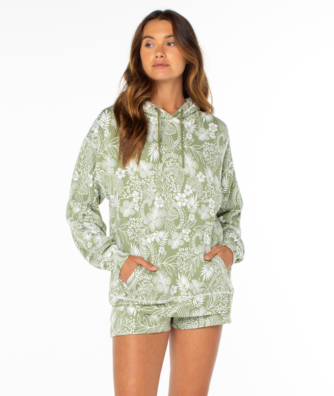 A woman wearing the Roxy Surfing By Moonlight Printed Hoodie in light green with white floral patterns stands with her hands in the hoodie pocket, looking slightly to the side against a plain white background.