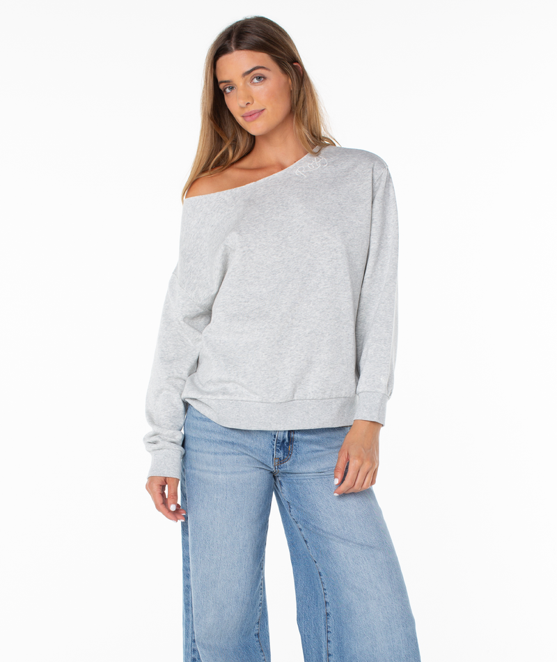 A woman with long hair wears Roxy’s Wave Break Top in Heather Grey Script—a luxe, off-the-shoulder sweatshirt crafted from a plush cotton blend—paired effortlessly with light blue jeans as she stands against a plain white background.