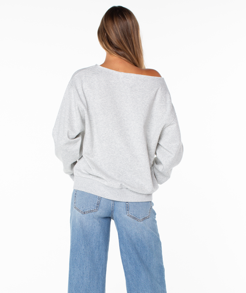 A person with long hair is standing with their back to the camera, wearing the Roxy Wave Break Top—a loose light gray plush cotton blend sweatshirt—and light blue jeans against a plain white background.