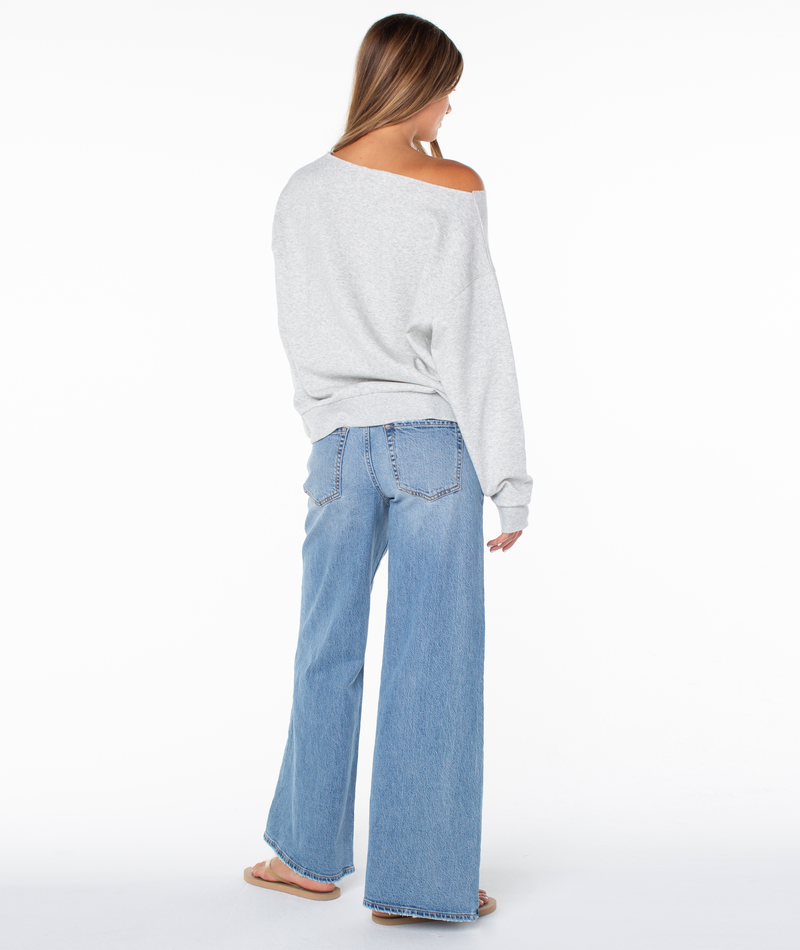 A woman stands facing away, wearing the Roxy Wave Break Top—an upscale, loose-fitting light gray off-the-shoulder sweatshirt crafted from a plush cotton blend. She pairs it with light blue wide-leg jeans and flip-flops. Her brown hair is down, set against a plain white background.