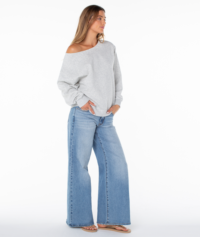 A woman stands against a white background, wearing an upscale loungewear look featuring the Wave Break Top by Roxy—a loose off-the-shoulder gray plush cotton blend sweater—paired with wide-leg blue jeans and sandals. She has her hands in her pockets and looks to the side.