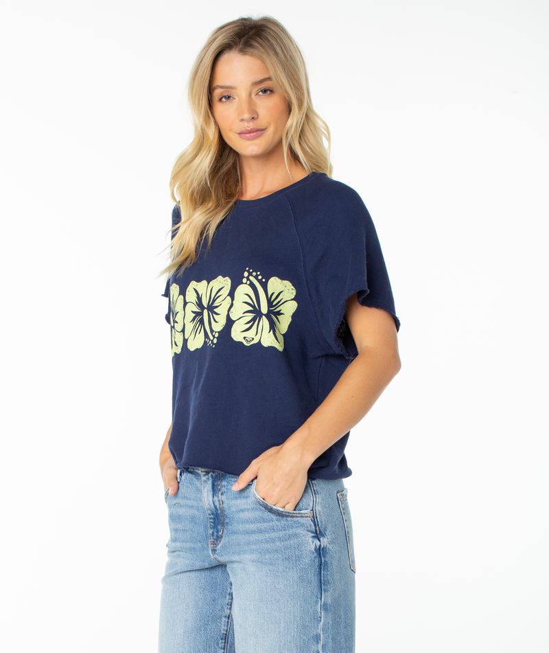 A woman with long blonde hair is wearing the Roxy Free Time Top in Naval Academy Flower Block—a cozy cotton t-shirt with yellow hibiscus flower prints, featuring a relaxed silhouette—paired with light blue jeans and standing against a white background.