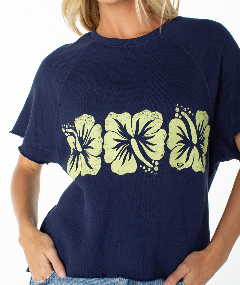 Wearing the Roxy Free Time Top, a navy blue cotton tee adorned with three yellow hibiscus flower prints across the front, this effortlessly casual piece features raw-edged short sleeves and a relaxed silhouette—perfectly styled with blue jeans.