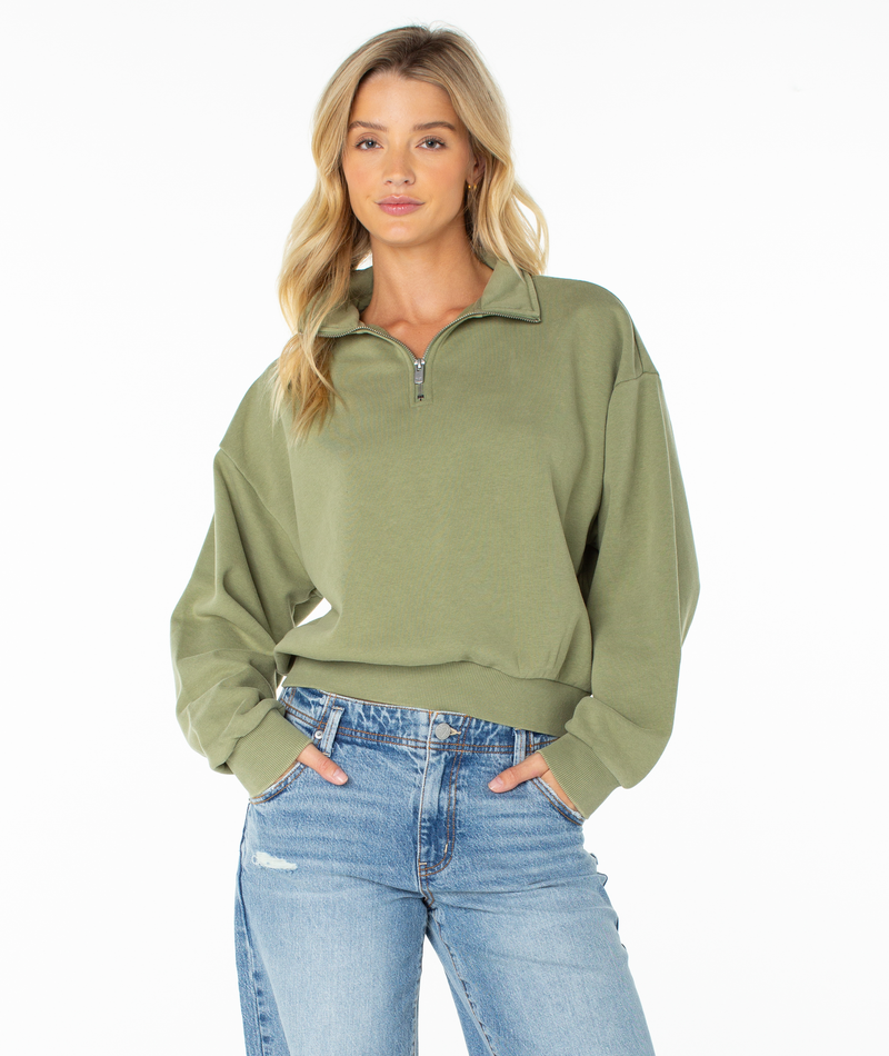 A woman with long blonde hair wears the Roxy Half Moon Top in Oil Green Solid, paired with light blue jeans, standing with her hands in her pockets against a plain white background.